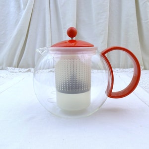 Puede incluir: Una tetera de vidrio transparente con tapa y asa rojas. La tetera tiene un filtro integrado y está diseñada para preparar té de hojas sueltas.