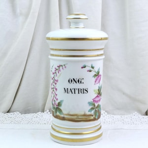 Puede incluir: Un tarro de farmacia de cerámica blanca con tapa, adornado con ribetes dorados. El tarro presenta ilustraciones florales en rosa y verde, y el texto "ONG: MATRIS" en negro. Artículo vintage.