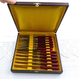 Könnte beinhalten: Ein Vintage-Besteckset in einer dunkelbraunen Box mit gelbem Samtinnenraum. Das Set enthält Gabeln und Messer mit dunkelroten Griffen und goldfarbenen Metallakzenten. Die Box ist geöffnet und zeigt das ordentlich angeordnete Besteck.