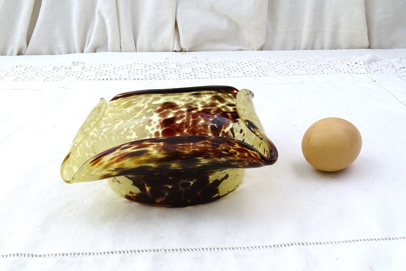 May include: A square-shaped glass bowl with a mottled brown and yellow pattern. The bowl has a unique, wavy design and is shown next to a brown egg.