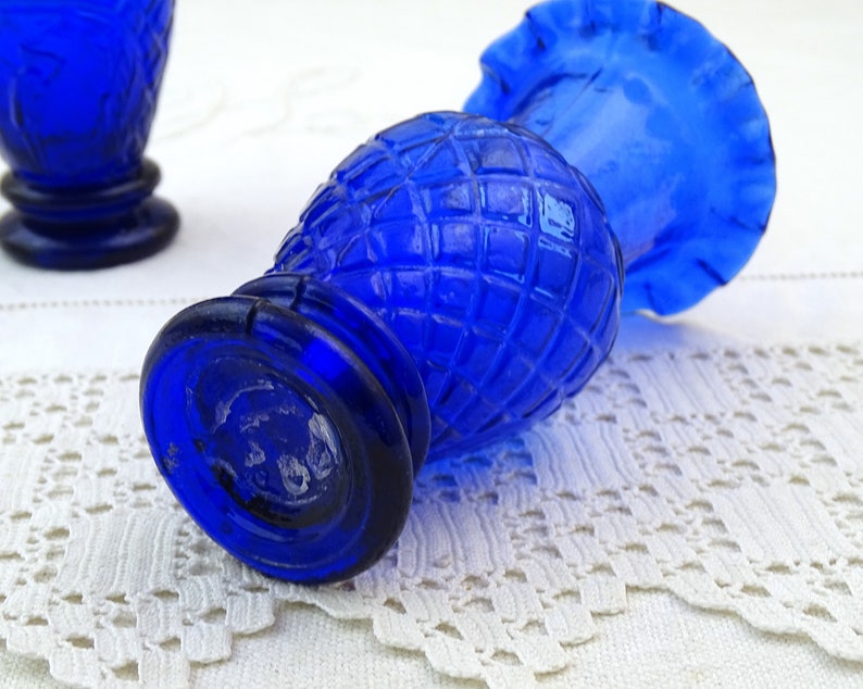 May include: Two cobalt blue glass bud vases with a diamond pattern and ruffled tops. The vases are sitting on a white crocheted surface.