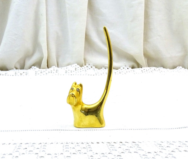May include: Gold-colored metal ring holder shaped like a dog with rhinestone eyes. The dog is sitting on a white lace surface.