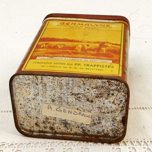 Puede incluir: Una lata rectangular y vintage de Germalyne con una etiqueta roja y amarilla. La etiqueta presenta texto y una ilustraci&oacute;n de personas cosechando trigo. La lata tiene un aspecto oxidado y el texto "LA MOISSON A L'ABBAYE DE SEPT-FONS" es visible.