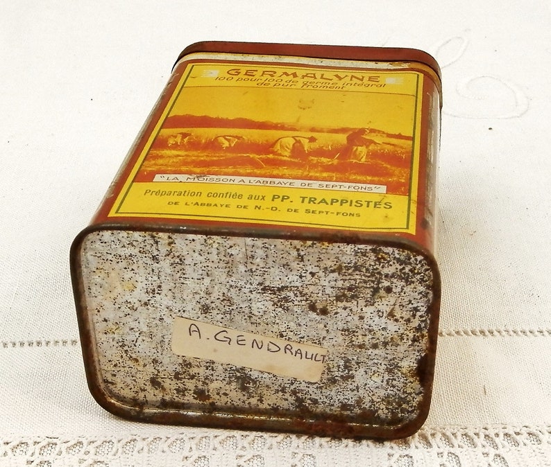 Puede incluir: Una lata rectangular y vintage de Germalyne con una etiqueta roja y amarilla. La etiqueta presenta texto y una ilustraci&oacute;n de personas cosechando trigo. La lata tiene un aspecto oxidado y el texto "LA MOISSON A L'ABBAYE DE SEPT-FONS" es visible.