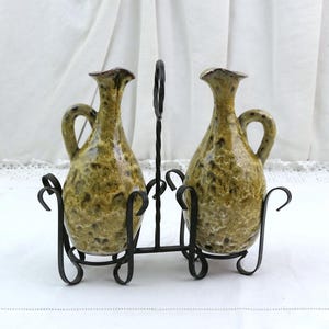 Può includere: Due ampolle in ceramica con una smaltatura maculata verde oliva e marrone, ciascuna dotata di collo stretto e manico laterale. Le ampolle sono inserite in un supporto in filo metallico nero con un anello centrale e supporti curvi per le bottiglie.