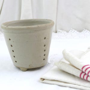 May include: A light beige ceramic berry bowl with a tapered shape, a thick rim, and small feet at the base. The sides of the bowl feature vertical rows of small, circular drainage holes evenly spaced around the exterior of the vessel.