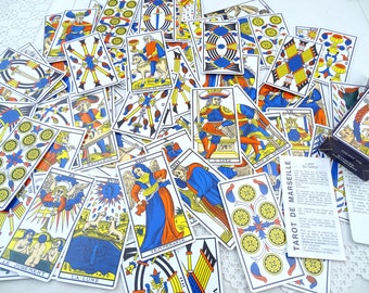 Puede incluir: Una baraja de cartas de Tarot de Marsella extendida sobre una superficie blanca. Las cartas son blancas con acentos azules, rojos y amarillos. Las cartas presentan varias imágenes y símbolos, incluyendo una espada, una copa, una varita y un pentagrama. La caja de las cartas es visible en la esquina inferior derecha de la imagen.
