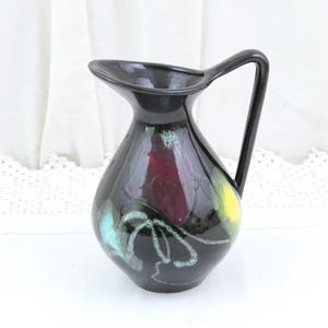 Può includere: Una brocca in ceramica nera con finitura lucida, dotata di un manico curvo e un ampio beccuccio. Il corpo è decorato con spruzzi astratti di rosso intenso, giallo brillante e blu turchese, accentuati da sottili linee bianche a spirale.