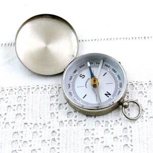 May include: A vintage silver compass with a lid, on a white lace background. The compass face is white with black markings and a blue needle. The word "GERMANY" is printed on the face.