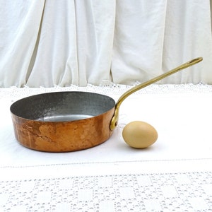 May include: A small copper saucepan with a hammered finish and a brass handle. The saucepan is sitting on a white lace tablecloth next to a brown egg.