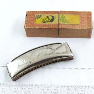 Può includere: Armonica argentata vintage con scatola marrone e gialla. L'armonica è curva e presenta una serie di fori. La scatola è di cartone e ha due etichette gialle con immagini. Gli oggetti sono su una superficie bianca.