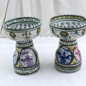 Può includere: Due portacandele in ceramica dipinti a mano. Ognuno ha una forma a ciotola ampia e poco profonda sopra una base a piedistallo. Le basi presentano disegni circolari colorati con motivi floreali e animali. I bordi sono decorati con motivi verdi e gialli.