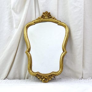 May include: An ornate gold-toned mirror with a scalloped frame. The mirror has a decorative design at the top and bottom of the frame.
