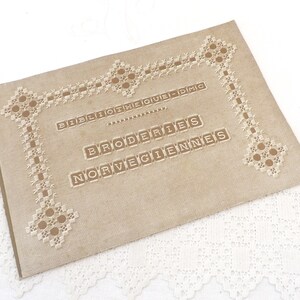 May include: A tan-colored book with a decorative border of white embroidery. The title of the book is "Bibliotheque DMC Broderies Norvegiennes".