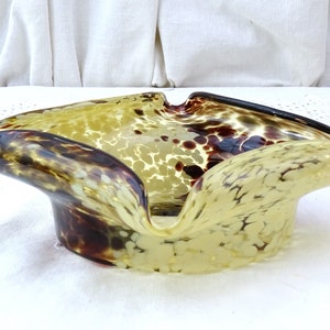 May include: A yellow and brown glass ashtray with a mottled, speckled pattern. The ashtray is shaped like a shallow bowl with a wide, rounded opening.