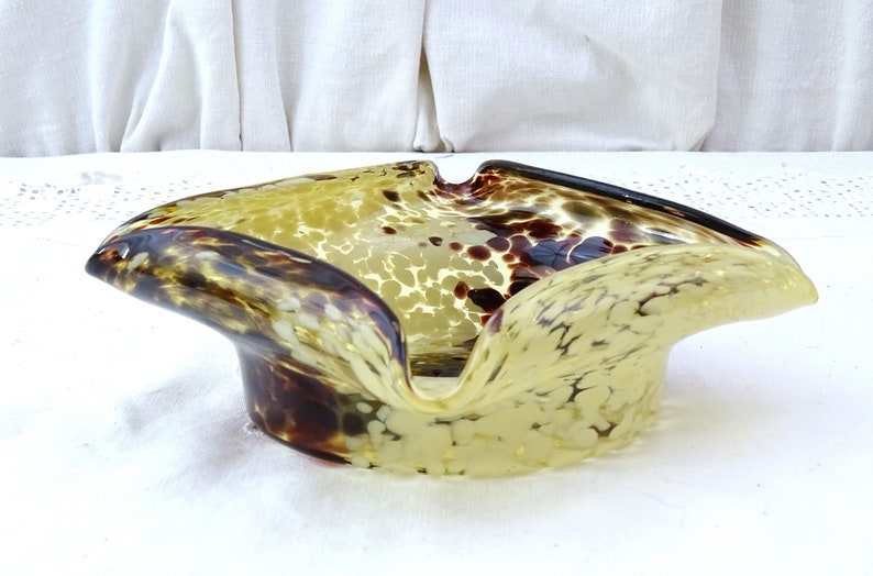 May include: A yellow and brown glass ashtray with a mottled, speckled pattern. The ashtray is shaped like a shallow bowl with a wide, rounded opening.