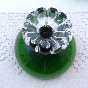 Puede incluir: Un tap&oacute;n de botella de vidrio verde con una parte superior en forma de flor de vidrio transparente. La flor tiene un centro negro.