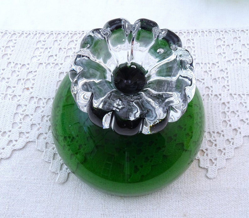 Puede incluir: Un tap&oacute;n de botella de vidrio verde con una parte superior en forma de flor de vidrio transparente. La flor tiene un centro negro.