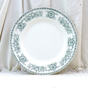 Può includere: Un piatto in ceramica bianca con un motivo floreale verde attorno al bordo. Il piatto è su una superficie bianca con un bordo in pizzo bianco.