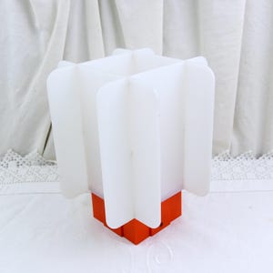 Pode incluir: Uma lâmpada geométrica branca e laranja. A lâmpada tem uma base quadrada na cor laranja brilhante. A parte superior é branca com quatro painéis verticais curvos. A lâmpada está sobre uma superfície branca.