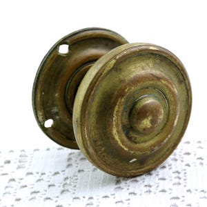 May include: Antique brass door knob set. The set includes two round knobs and a backplate. The knobs have a circular design with a central button. The brass has a weathered appearance with areas of discoloration, suggesting age and use. The backplate has two screw holes.