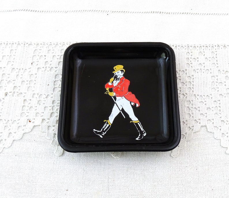 May include: Black square ashtray with a white and red image of a man in a red jacket and a black top hat walking with a stick. The man is wearing white pants and black boots.