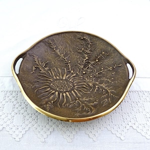 以下が含まれることがあります： ブロンズ色の装飾トレイ。花柄のデザインで、浮き彫りのヒマワリと植物の要素が特徴です。 金色の縁と2つのハンドルが付いています。 白いテクスチャのある表面に置かれています。