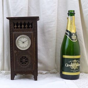 May include: A small wooden clock with a carved door and a white face. The clock is standing on a white surface next to a bottle of champagne.