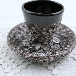 Può includere: Un portauovo in ceramica nera con base e piattino strutturati e maculati. Il bordo della tazza è liscio e lucido, in contrasto con l'aspetto ruvido e simile alla pietra della base e del piattino. Il set è progettato per servire le uova.