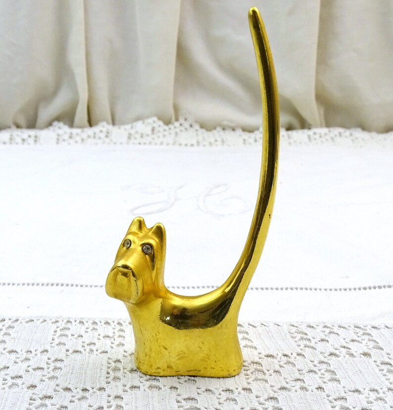 May include: Gold-colored ring holder shaped like a dog with rhinestone eyes. The dog is sitting up with its tail curled around its back.