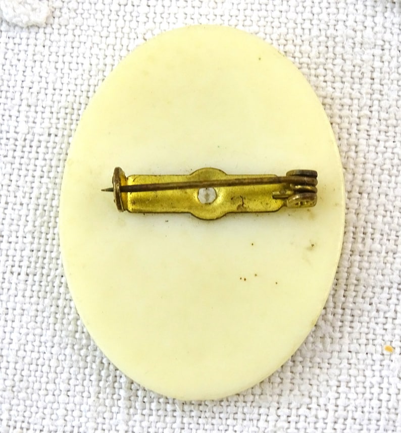 May include: The back of a white round pin with a gold metal pin back.