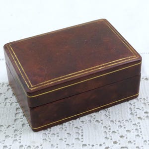 May include: A rectangular brown box with a hinged lid. The box has gold trim around the edges. The box is sitting on a white lace tablecloth.