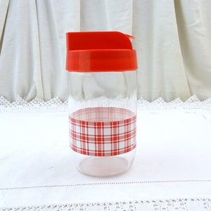 May include: A clear glass jar with a red lid and a red and white plaid pattern around the middle. The jar is sitting on a white tablecloth.