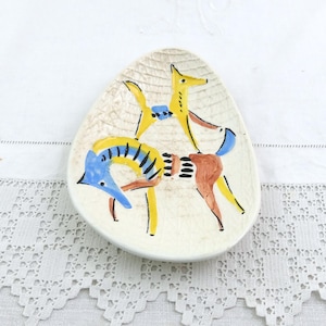 Puede incluir: Un pequeño plato de cerámica blanquecina con superficie texturizada. El plato presenta un diseño abstracto y colorido de dos animales estilizados en amarillo, azul y marrón. Los animales parecen estar interactuando. El plato está sobre un mantel blanco de encaje.