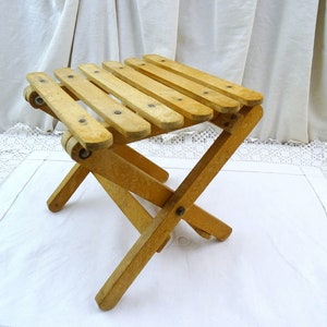 May include: A wooden folding stool with a light brown finish. The stool has a simple design with a rectangular seat and four legs that fold up for easy storage.