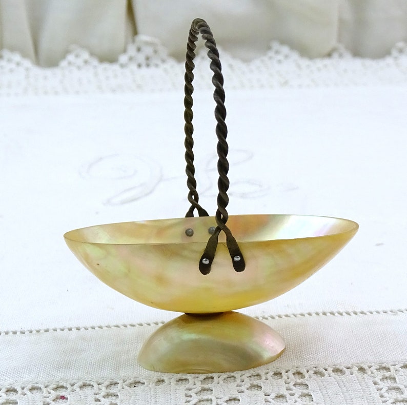 May include: A small, oval-shaped bowl made of iridescent mother-of-pearl with a twisted wire handle. The bowl is on a white surface.