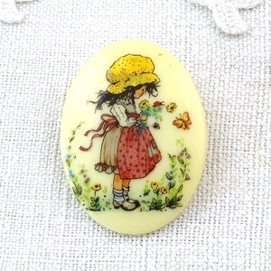May include: A vintage oval-shaped button with a yellow background featuring a girl in a yellow hat and red and white dress holding a bouquet of flowers. The girl is surrounded by flowers and butterflies.