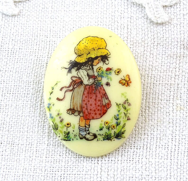 May include: A vintage oval-shaped button with a yellow background featuring a girl in a yellow hat and red and white dress holding a bouquet of flowers. The girl is surrounded by flowers and butterflies.