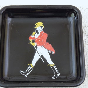 May include: Black square ashtray with a raised rim. A man in a red jacket, white pants, and a yellow hat is walking with a cane.
