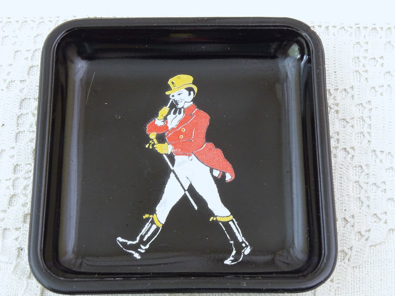 May include: Black square ashtray with a raised rim. A man in a red jacket, white pants, and a yellow hat is walking with a cane.