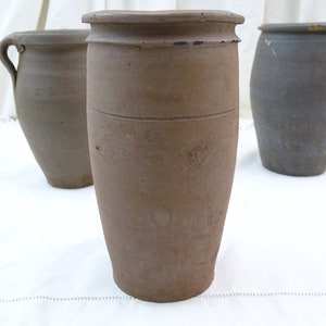 May include: Three brown ceramic pottery jars, two with wide mouths and one with a narrow mouth. The jars are on a white tablecloth.