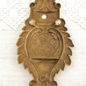 May include: A brass wall mount with a decorative, ornate design. The mount has a rectangular opening in the center and a raised, floral design around the edges.