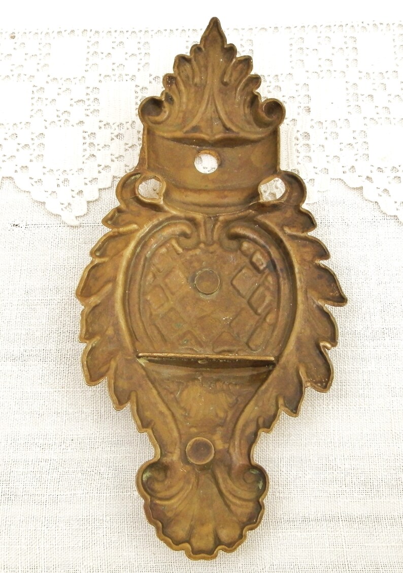 May include: A brass wall mount with a decorative, ornate design. The mount has a rectangular opening in the center and a raised, floral design around the edges.