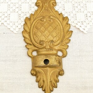 May include: A gold-toned metal decorative wall bracket with a ornate design. The bracket has a central oval shape with a criss-cross pattern and a small hole in the center.