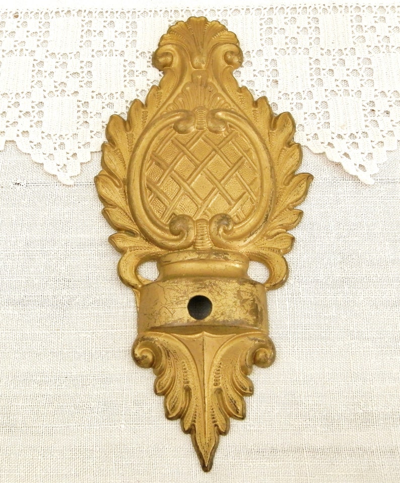 May include: A gold-toned metal decorative wall bracket with a ornate design. The bracket has a central oval shape with a criss-cross pattern and a small hole in the center.