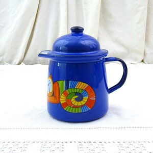 Puede incluir: Una cafetera de esmalte azul cobalto con asa y tapa negras. La cafetera presenta un diseño caprichoso de gato naranja con una cola en espiral colorida. La cafetera está sobre una superficie blanca.