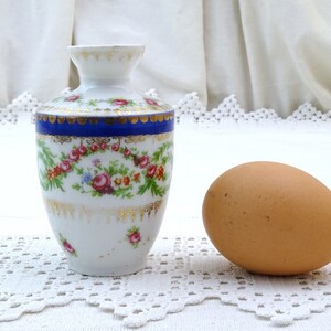 May include: A small white porcelain vase with a blue and gold trim and floral pattern. The vase is decorated with pink, red, and yellow flowers. The vase is sitting on a white lace tablecloth next to a brown egg.