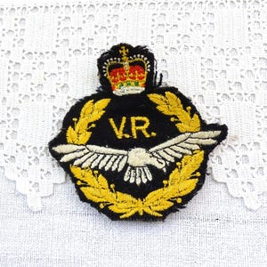 May include: Embroidered patch featuring a black background with gold laurel leaves, a crown, and the letters "V.R." in gold. A white bird with outstretched wings is centered below the letters. The patch is likely a vintage military or commemorative item.