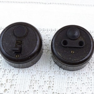 May include: Two vintage black bakelite electrical switches. One is a light switch with a toggle and the other is an outlet with two round holes.