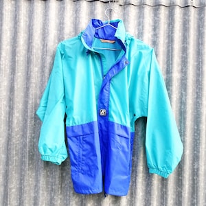May include: A vintage turquoise and blue windbreaker jacket with a hood and front pockets. The jacket is hanging on a metal hanger against a corrugated metal background. The jacket has a zipper closure and a logo on the chest.
